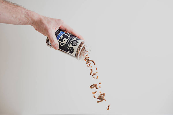 A hand holding a bottle of Beef Sprinkles with a sprinkling of beef powder coming out of it against a white background.