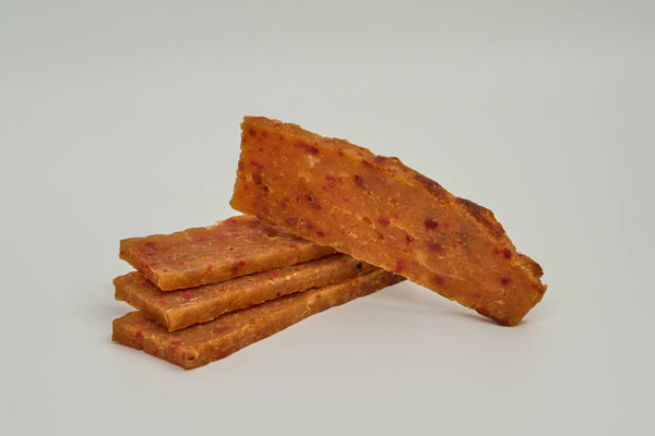 Four rectangular pieces of spicy meat jerky are stacked on a light surface, with one piece leaning diagonally on top. The jerky has a reddish-brown color with visible seasoning specks.