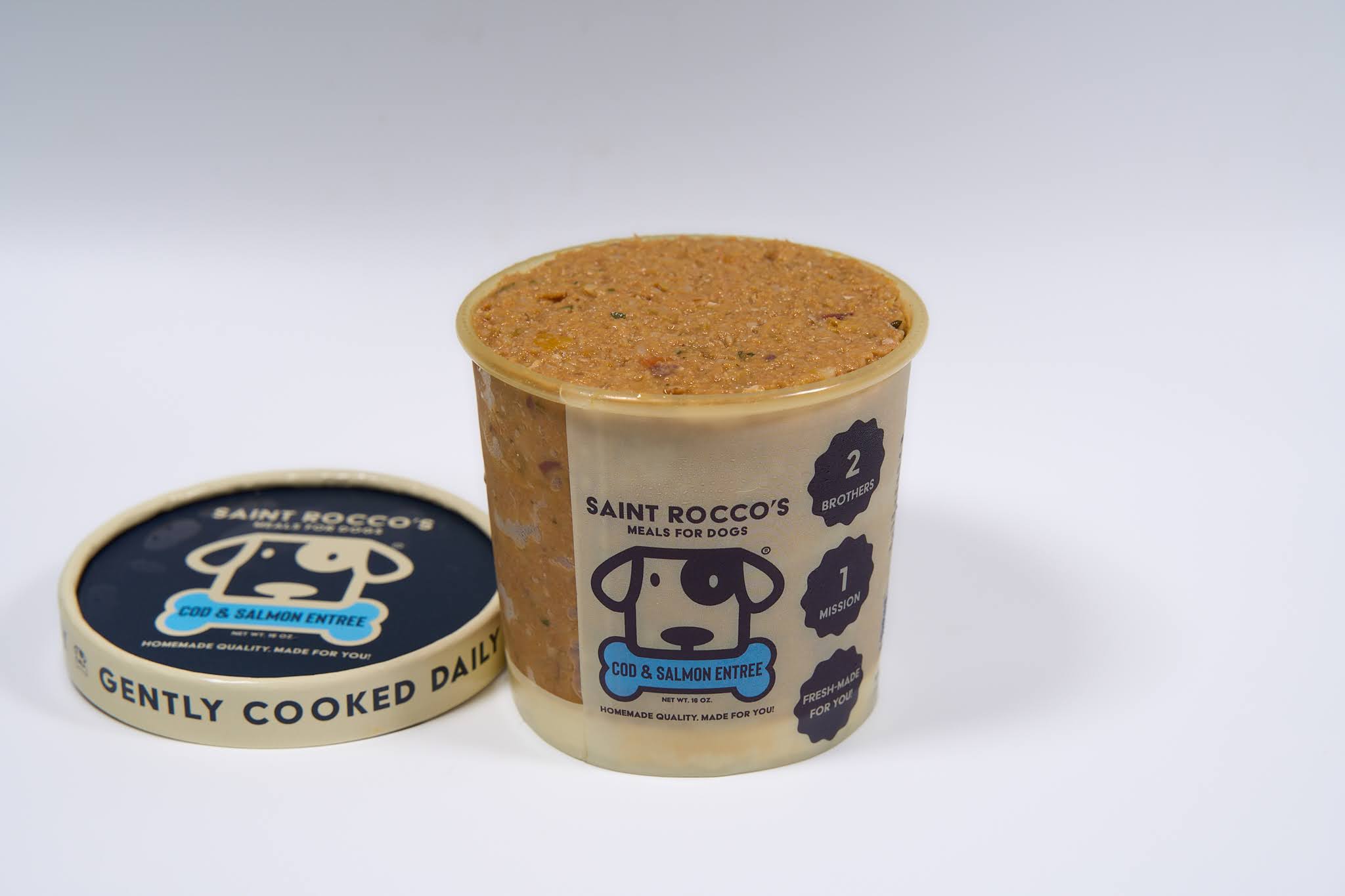 A container of Saint Rocco's Cod & Salmon Entree by Saint Rocco's Treats sits on a white background with its lid beside it, packed full and featuring a cartoon dog face and clear feeding instructions on the packaging.