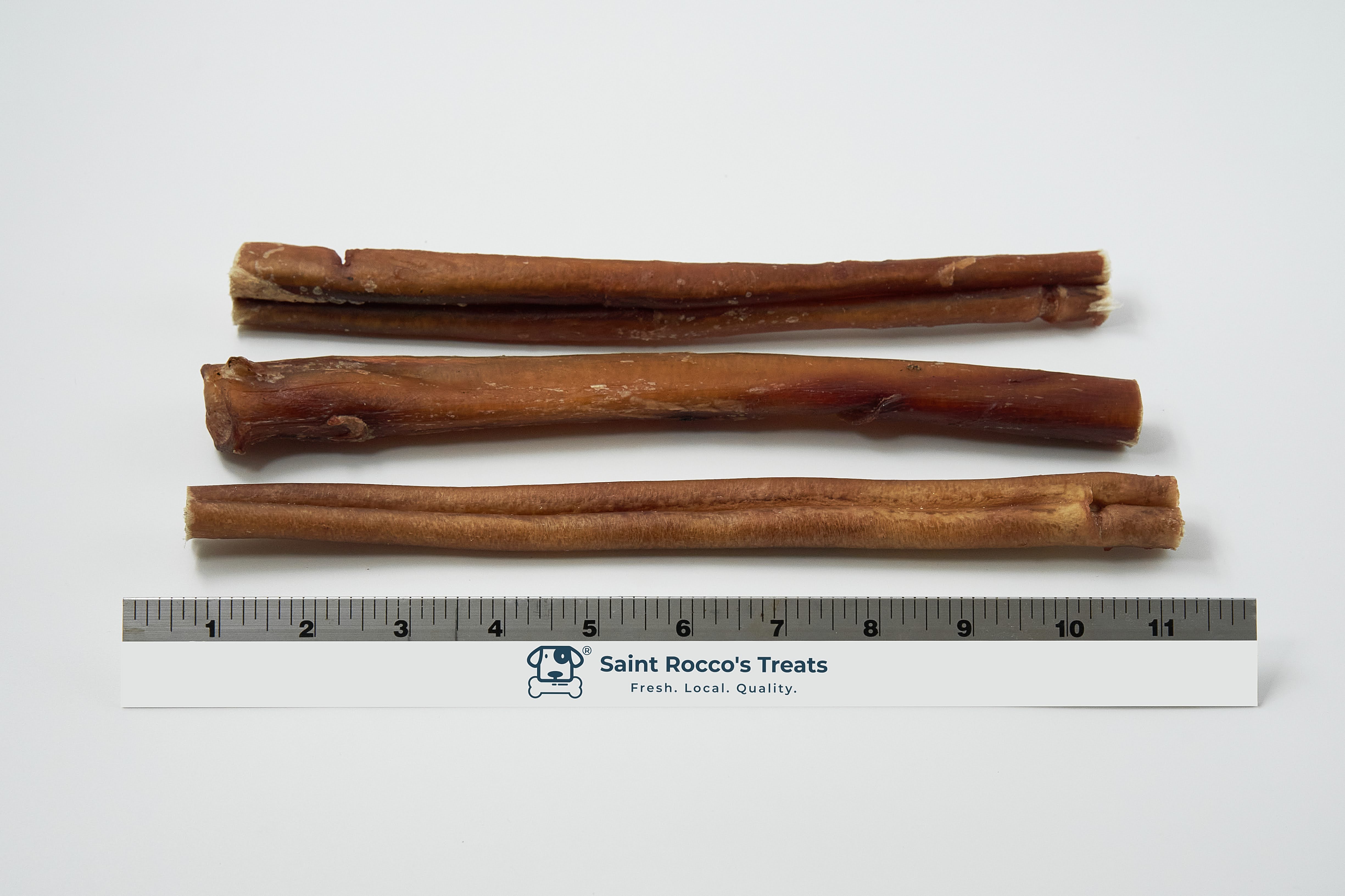 Three Saint Rocco's Bully Sticks of varying thicknesses are arranged parallel above a 15-cm Saint Rocco's Treats ruler. These all-natural dog chews are shown on a white background.