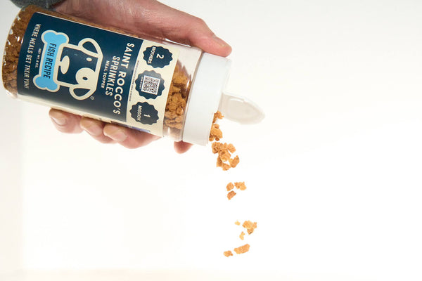 A hand holding a bottle of 'Saint Rocco's Promise' dog treat sprinkles, with the sprinkles visible in the process of being poured out.