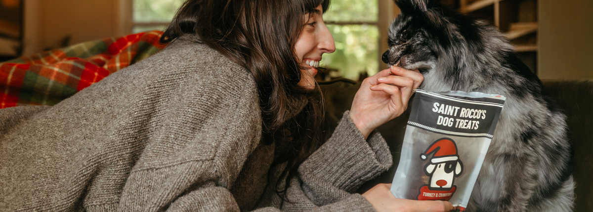 A woman in a cozy knit sweater smiles while holding a Saint Roch's Dog Treats bag next to a fluffy black and white dog in a warm indoor setting.