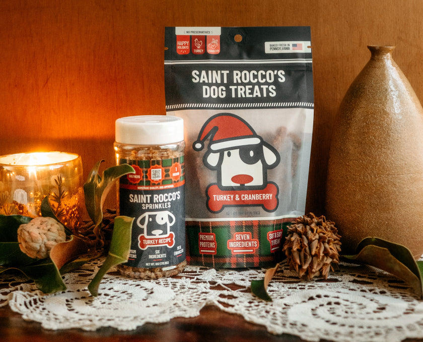 Saint Rocco's dog treats displayed on a table with a warm holiday setting, including a bag of Turkey & Cranberry treats and a jar of Spinkles, styled with a candle, lace doily, pinecones, and a ceramic vase.