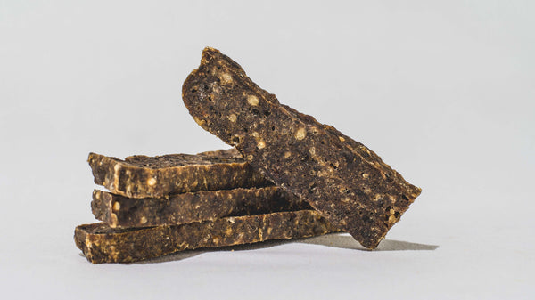 Four rectangular, brown Saint Rocco's Treats Training Bundle dog treats with visible seeds are stacked unevenly on a light surface, one bar leaning diagonally atop the pile against a plain white background.