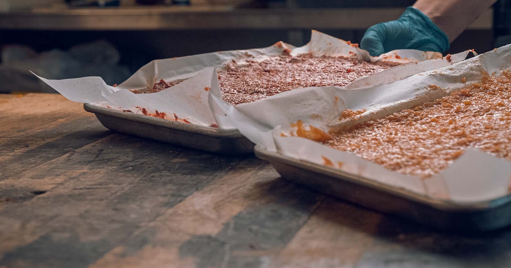 Two metal baking trays lined with parchment paper hold a baked, golden-brown dessert. A person wearing a blue glove is adjusting one tray on a wooden countertop in a kitchen setting.