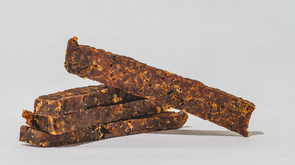 Three pieces of Saint Rocco's Treats Specialty Bundle, high-value brown beef jerky dog treats, are stacked on a plain white surface, with one piece resting diagonally on top of the others.