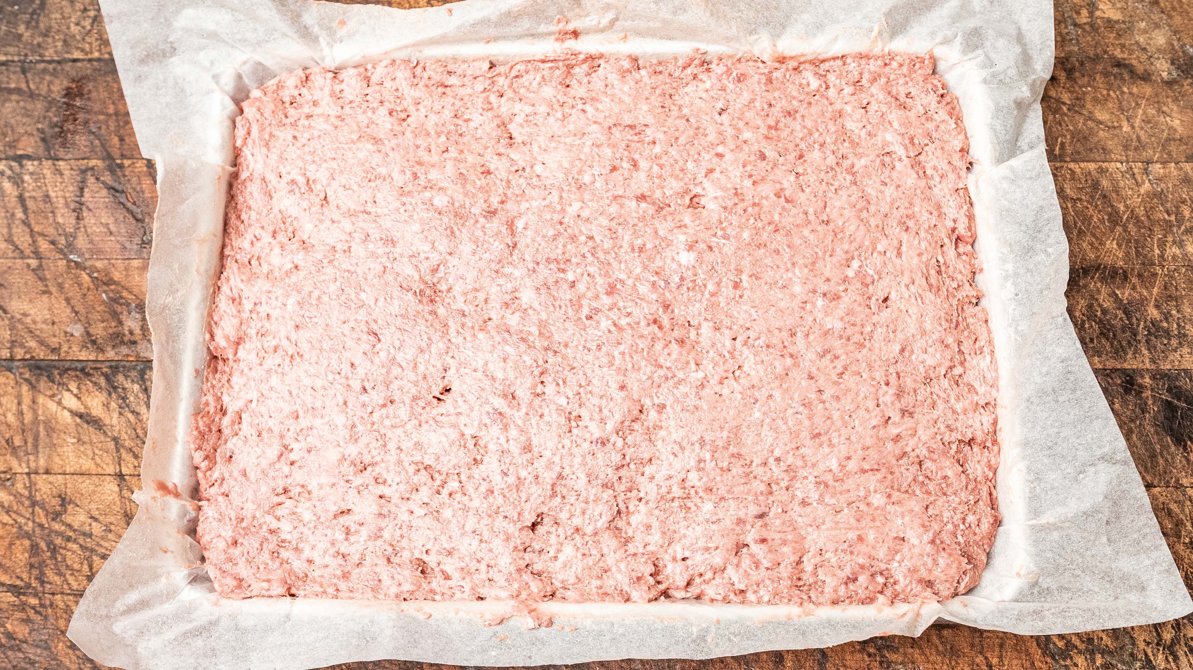 A rectangular baking pan lined with parchment paper is filled with an even layer of raw ground meat mixture, ready to be cooked, placed on a rustic wooden surface.