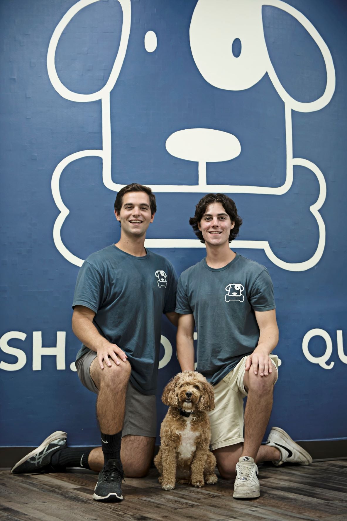 Two individuals and a dog sitting in front of a blue wall with a large white graphic of a dog's head