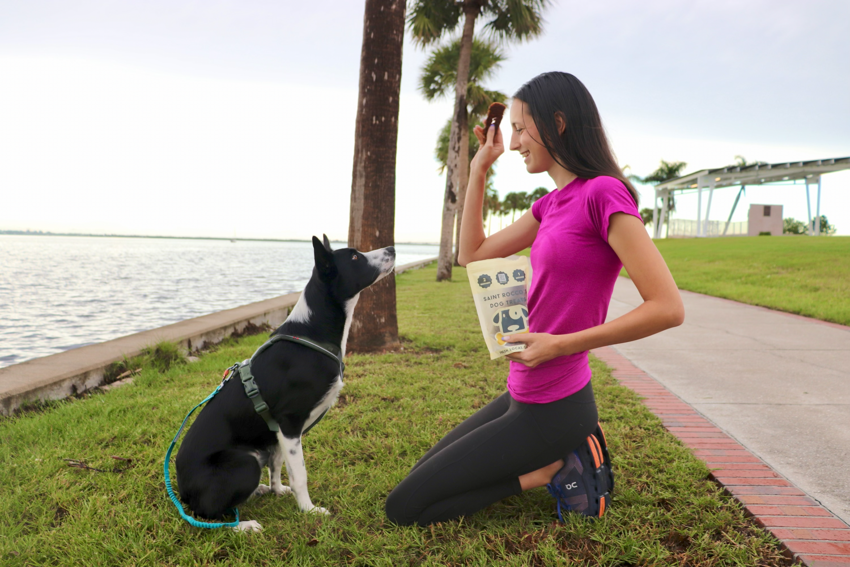 Woman in pink shirt and black leggings kneeling on grass with a dog by a waterfront path.
