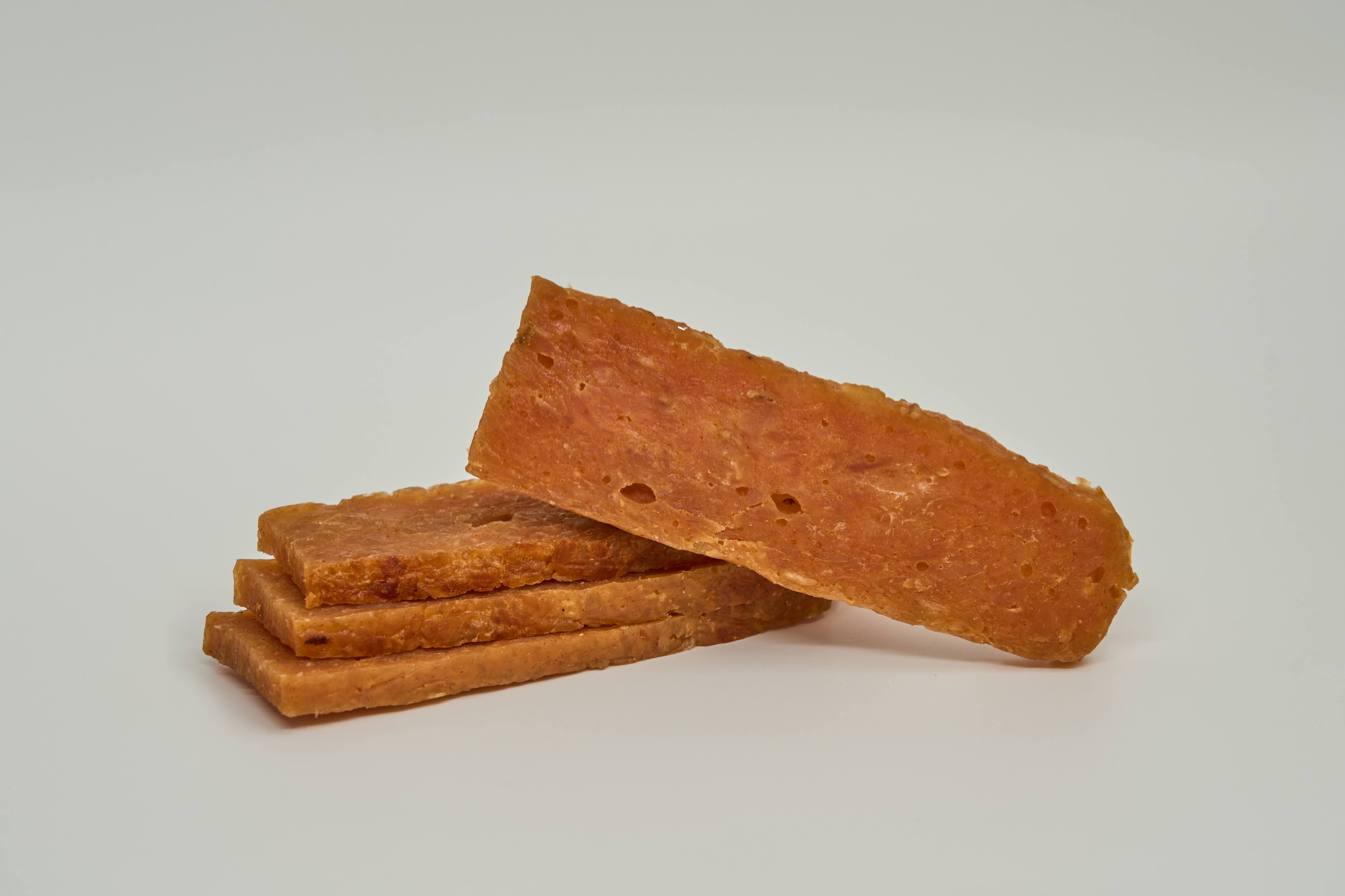 Four rectangular slices of Saint Rocco's Treats Chicken and Sweets Treats are stacked on a light surface: three lay flat, while one stands upright to display its textured reddish-brown surface.
