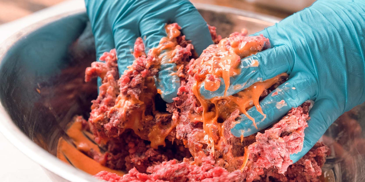 Hands wearing blue gloves mix raw ground meat and a yellow-orange liquid, possibly egg or sauce, in a large metal bowl.