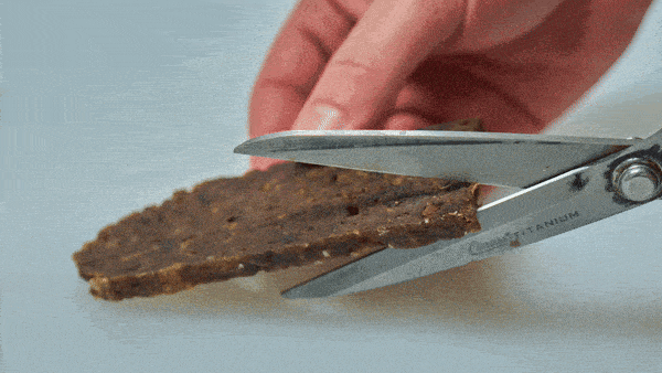 A hand uses scissors to cut a piece of brown artisanal dog jerky from the Saint Rocco's Treats Specialty Treats Bundle on a white surface.