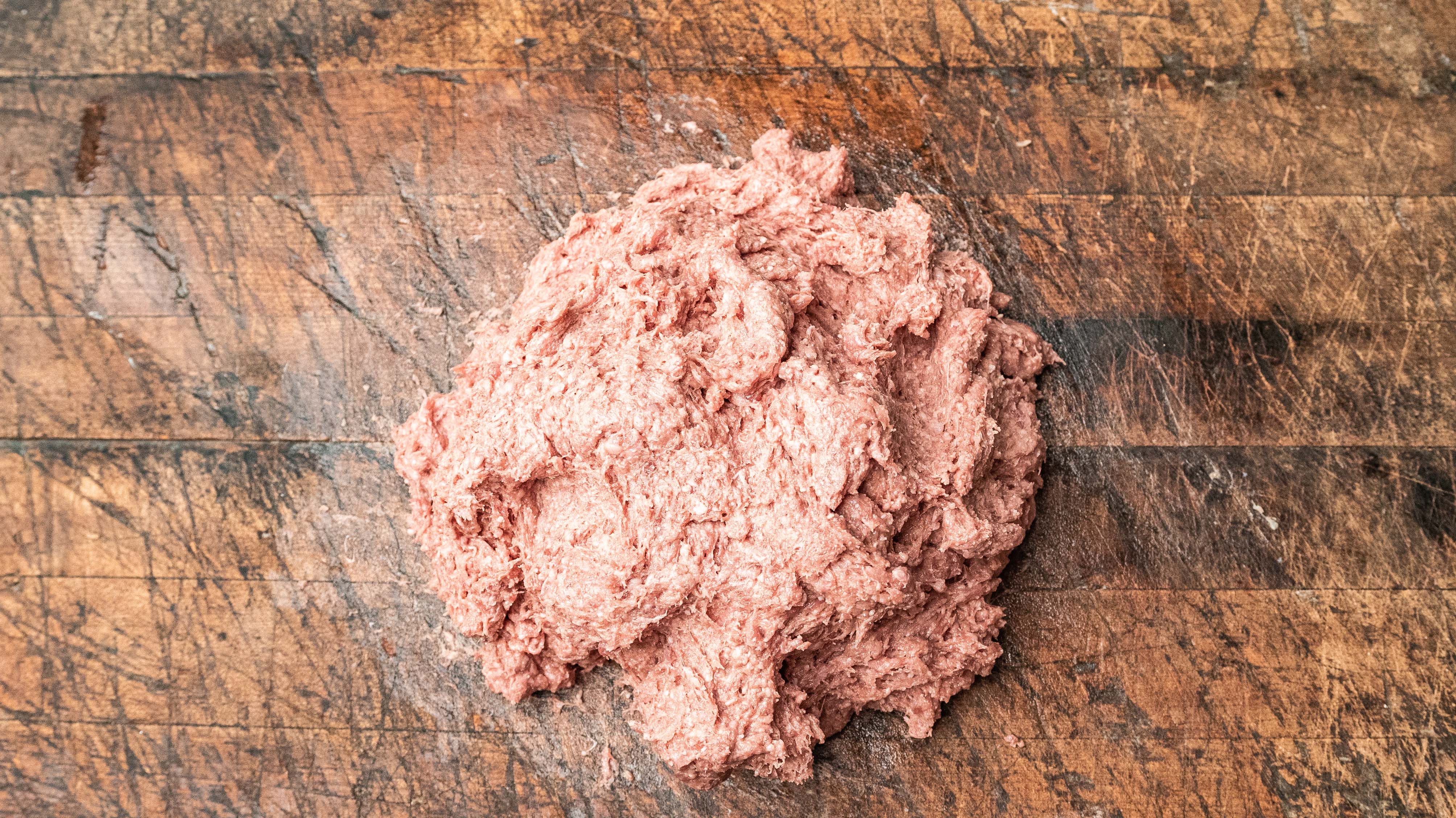 A mound of raw ground meat is placed on a worn wooden cutting board with visible scratches and knife marks.