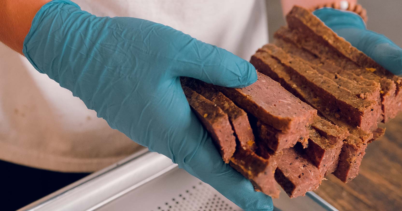 A person wearing blue gloves holds several thick slices of brown, loaf-like food above a metal tray or counter.