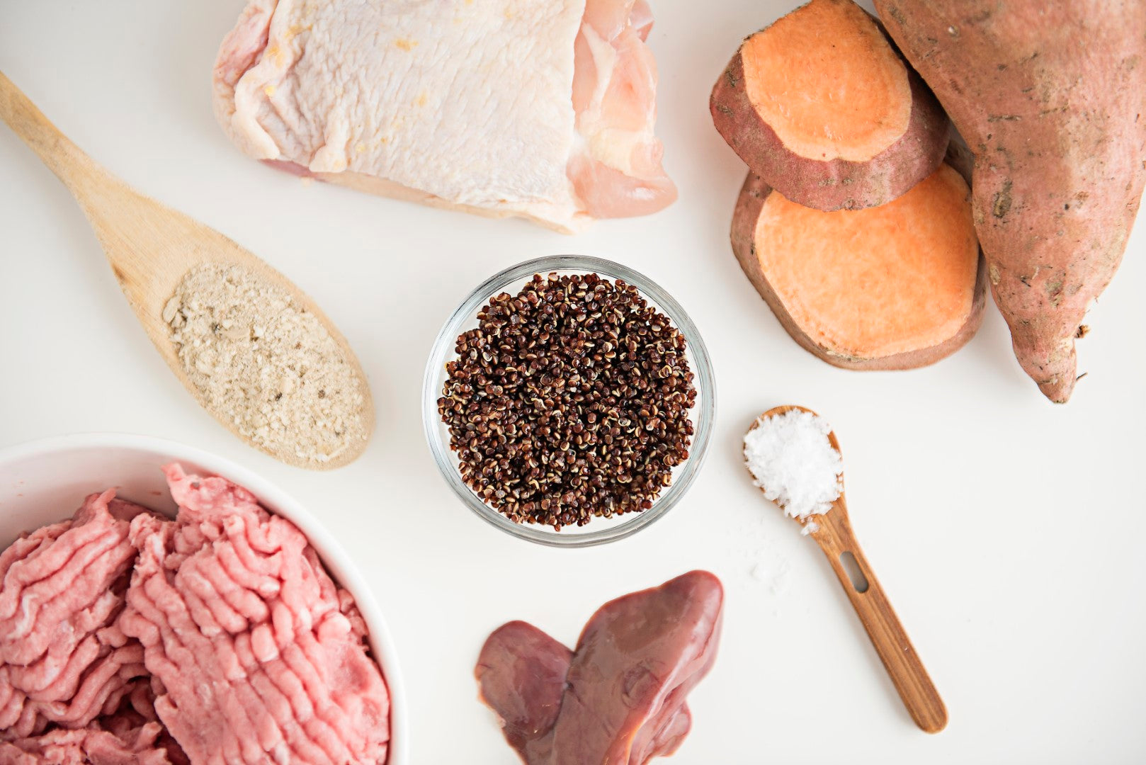 A flat lay of Entree Sampler ingredients by Saint Rocco's Treats on a white surface, including ground meat, raw chicken thigh, sweet potatoes, quinoa, chicken heart, and flour and salt on wooden spoons.